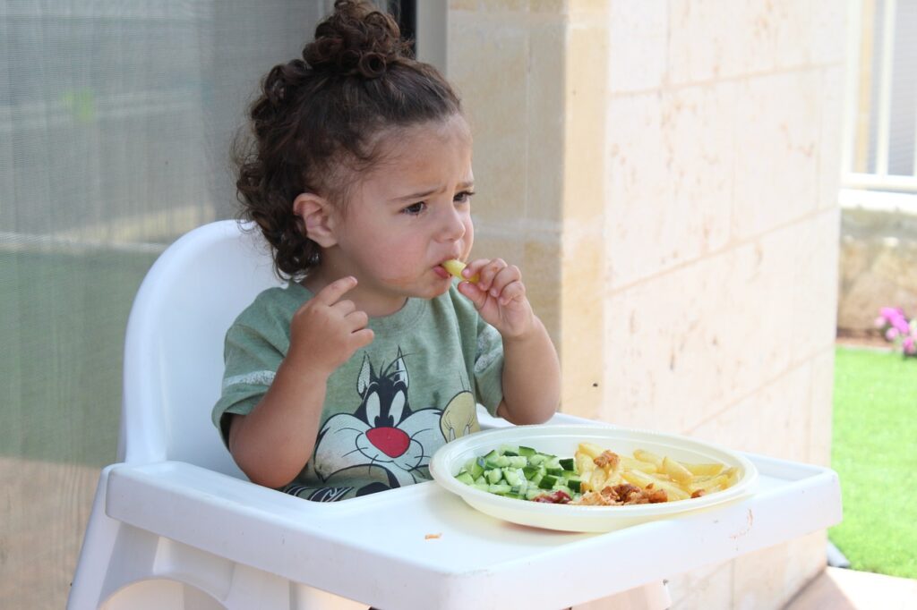 A toddler in a high chair eating pasta with a troubled facial expression