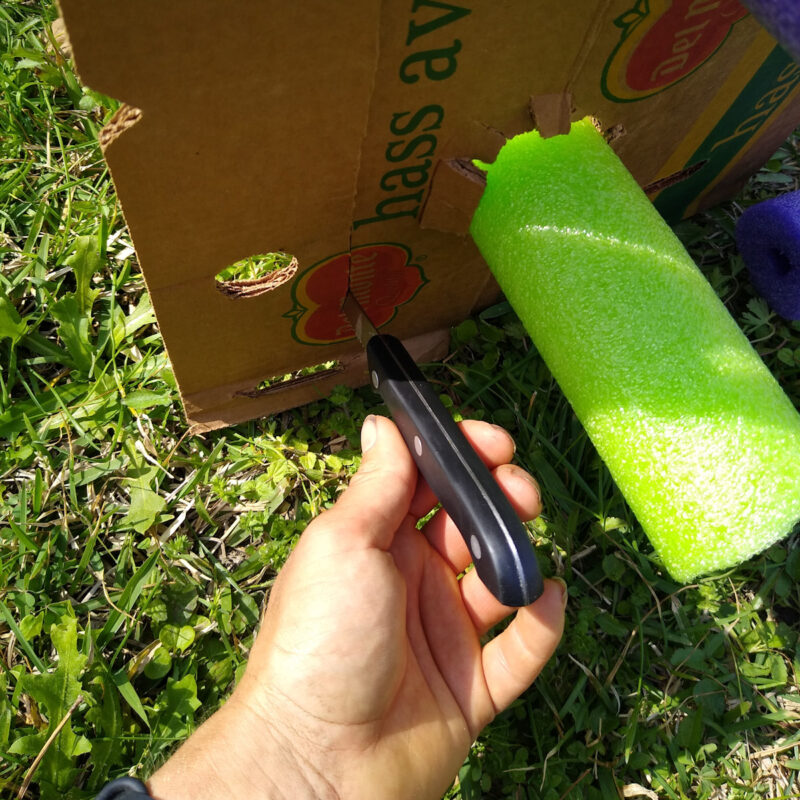 Close up of serrated knife cutting top flaps off of cardboard box. To right, green pool noodle sticks out of box.