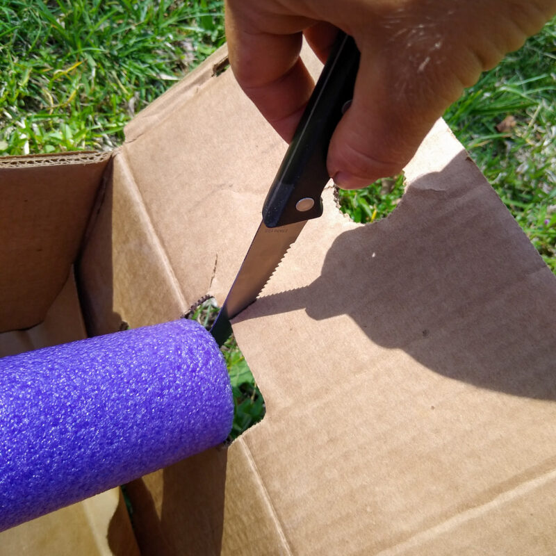 A serrated knife makes a hole in a cardboard box for a purple pool noodle to fit through.