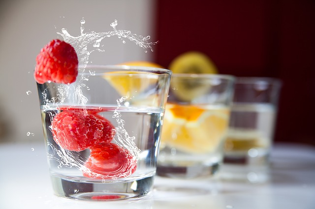 In foreground, a small glass of water. Two red raspberries in the glass, a third appears to be bouncing from the glass with splashing water surrounding.