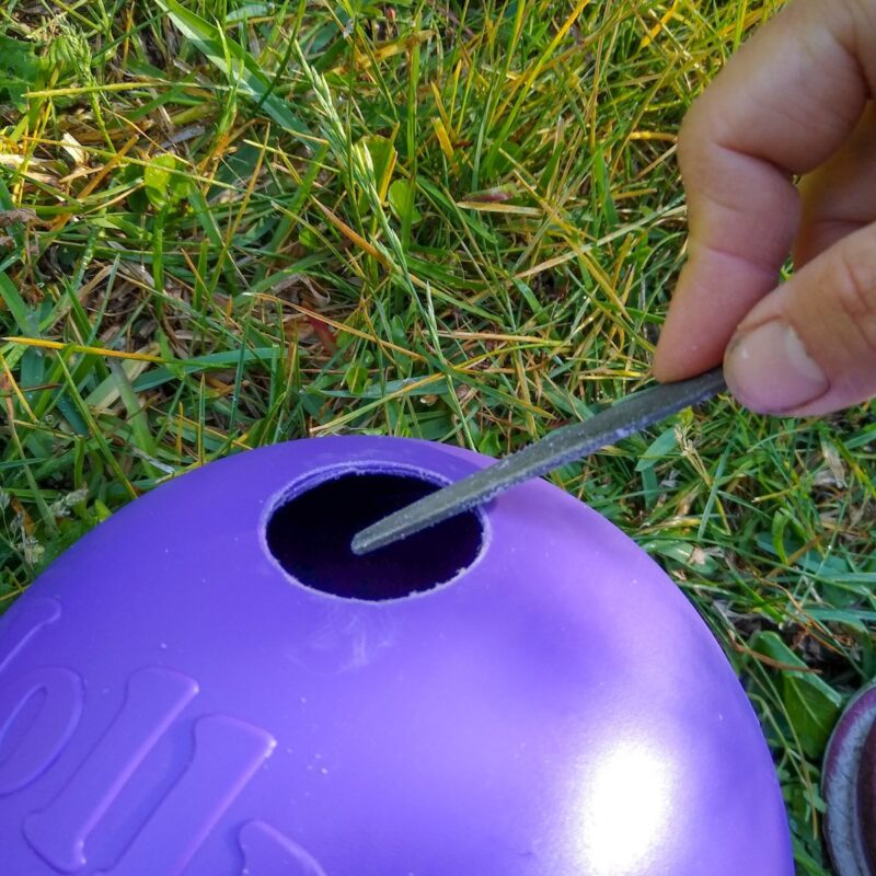 A close shot of a purple Jolly Egg with a hole cut with a hole saw. To the right a hand holds a small file and is removing plastic threads from the hole.