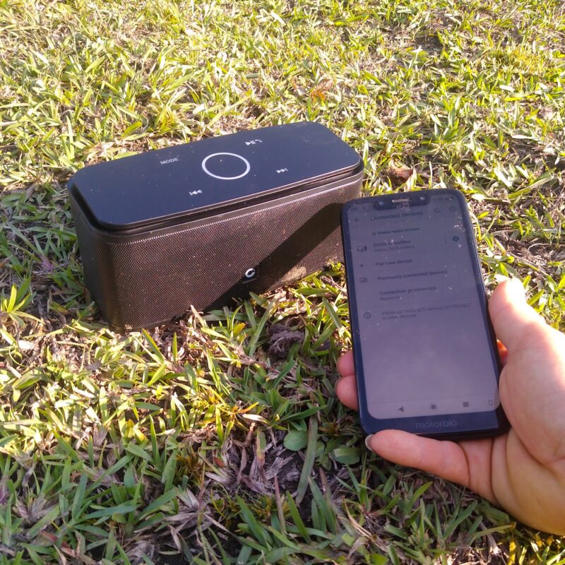 A speaker in a pasture on a grass background. A hand holding a mobile phone in foreground with music for horses about to play.
