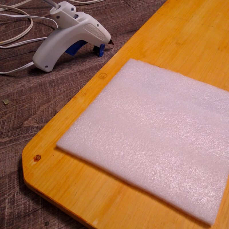 The wooden texture board with a square of soft white foam ready to be glued. A hot glue gun in background.