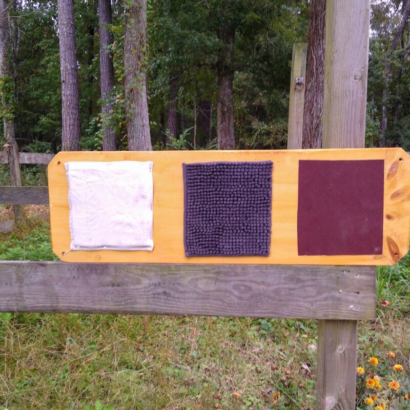 A completed horse stall enrichment texture board with three textures, resting on a fence. From left to right are canvas, gray chenille, and red sandpaper.