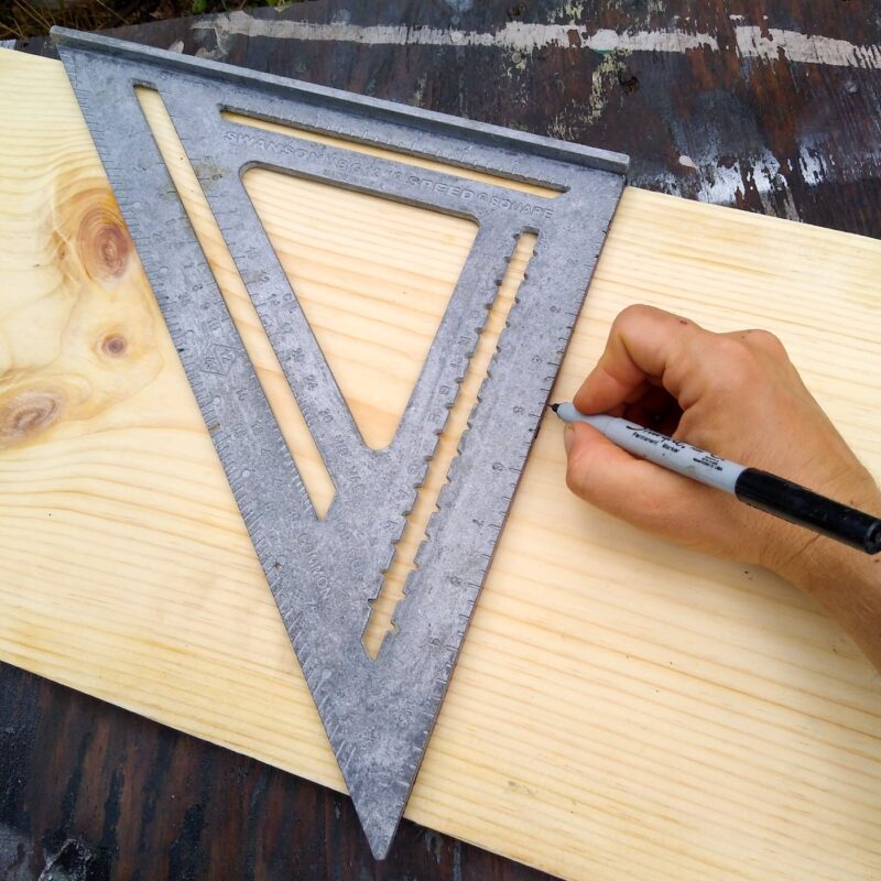A carpenters square on a pine wood board and hand marking a line for cutting.