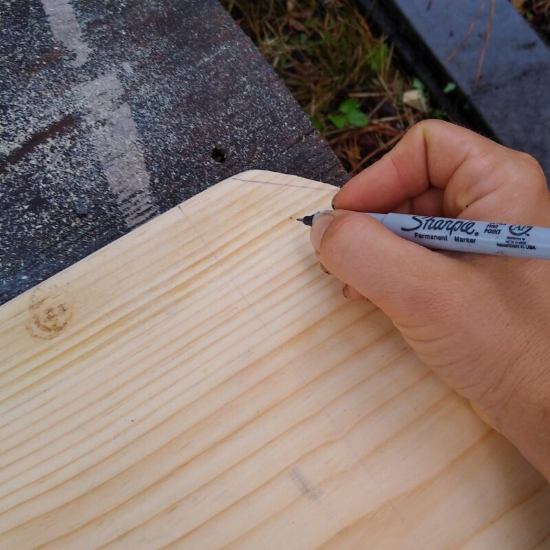 A hand with pen on the unfinished wooden enrichment board marking pilot hole locations. 
