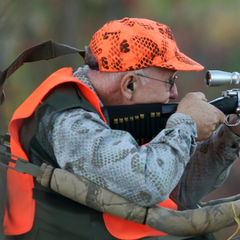 An older man in hunting equipment and orange safety hat and vest seen in profile looking into the sight of a rifle.