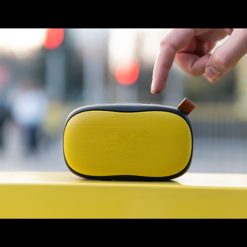 A yellow wireless speaker with a finger extending from top frame about to press a button. Blurry urban background.