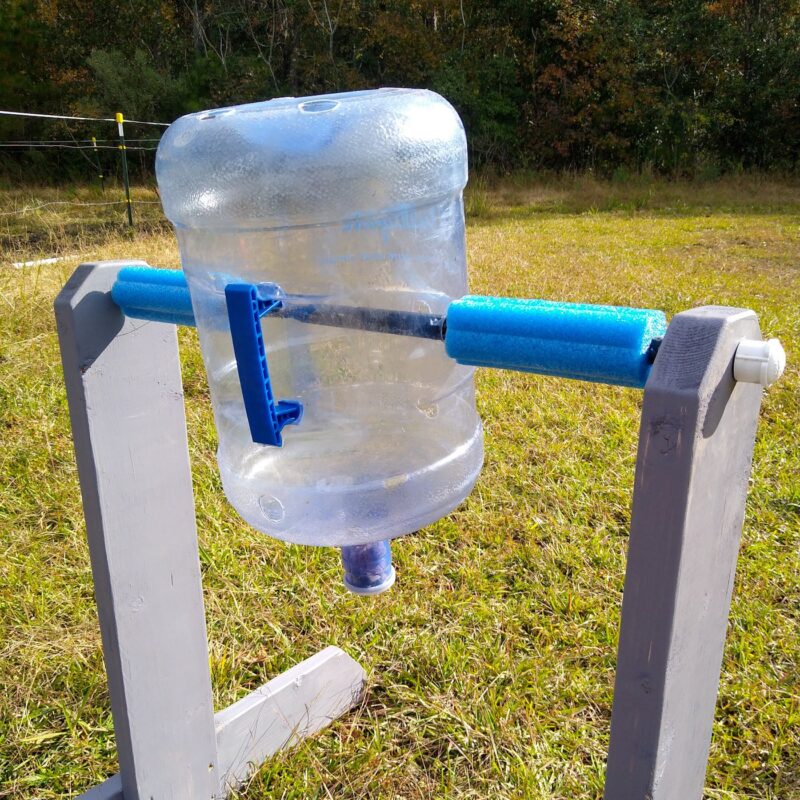A gray painted spin the bottle horse toy with five gallon water bottle attached and blue pool noodles covering the axle rod.