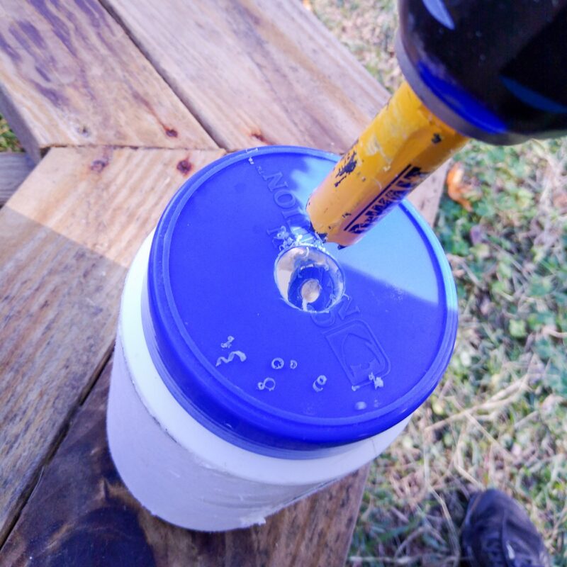 A blue plastic jar lid with yellow hole saw drill attachment making a hole in the lid.