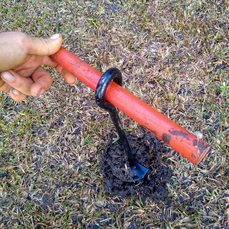 A hurricane tie or ground anchor seen from above, with piece of red painted metal pipe inserted into hay net anchor hole for leverage.