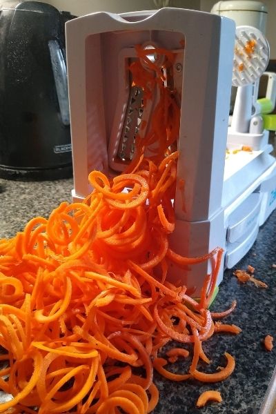 A hand crank veggie noodle maker or spiralizer shown with a pile of freshly cut carrot noodles.