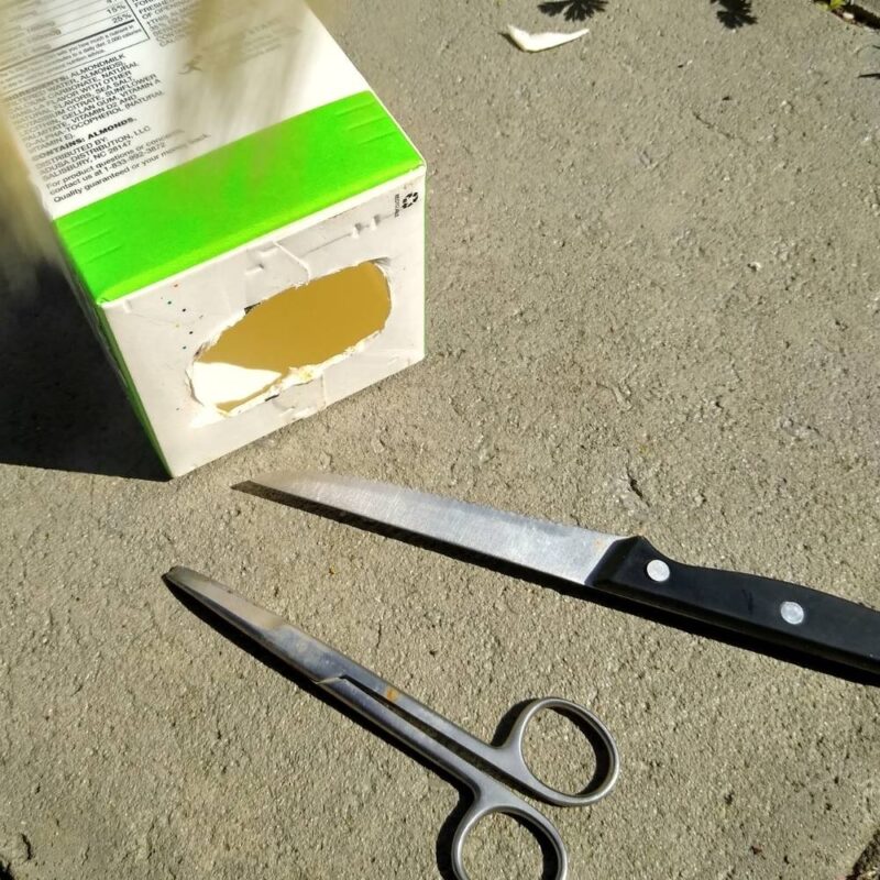 Almondmilk carton on side with hole cut in bottom. In foreground, serrated knife and scissors.