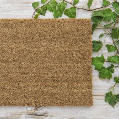A coconut fiber mat on a wooden floor with ivy in upper right corner.
