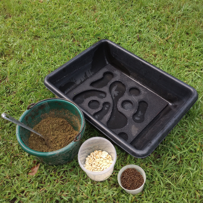 DIY horse stall mat slow feeder in a black plastic tray on green grass background. Horse feed and treats in buckets in foreground.