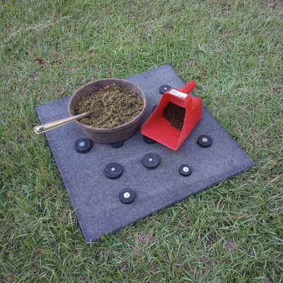 DIY horse slow feed mat rests on grass background. On top of mat is a round rubber pan of soaked hay pellets and a red feed scoop of dry horse feed.