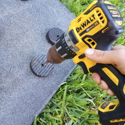 A yellow and black DeWalt power drill makes a hole in round rubber insert and rubber base mat for DIY horse slow feed mat.