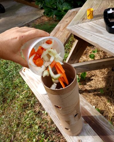 A hand from left of image holds a container of carrot and celery sticks being poured into the DIY cardboard horse toy against a picnic table background.