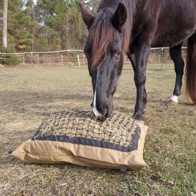 A black horse in background eats from a full Hay Pillow in foreground.